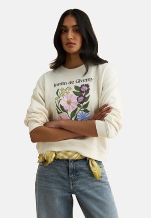 Donna con lunghi capelli scuri che indossa una felpa bianca con stampa floreale colorata e la scritta "Jardin de Giverny", jeans blu e una sciarpa gialla legata in vita.