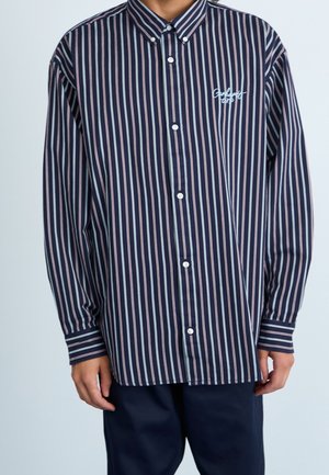 Osoba ubrana w granatową koszulę na guziki z pionowymi białymi i różowymi paskami oraz z haftem "Godland Corp" na piersi.