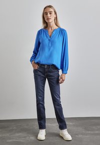 Blå långärmad blus med V-ringning och rynkade axlar, ihopparad med mörka jeans och vita sneakers på en grå bakgrund.