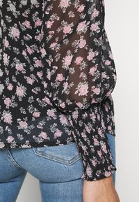Black floral blouse with pink roses; sheer fabric; puffed sleeves; gathered cuffs; paired with fitted blue jeans.