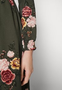 Gros plan d'une main reposant à côté d'une manche de robe vert foncé à motif floral avec des fleurs roses, jaunes et rouges.