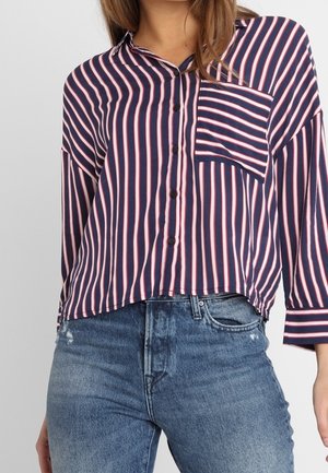 Gestreifte Button-Down-Bluse in Marineblau und Pink mit Kragen, Brusttasche und lockerer Passform, kombiniert mit hellblauen, distressierten Jeans.