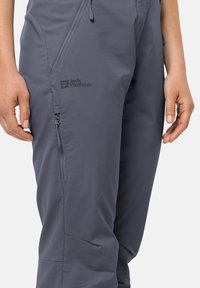 Grey outdoor pants made of softshell material, featuring a zipper pocket and the "Jack Wolfskin" logo on the thigh. Lightweight and durable design.