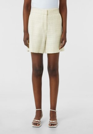 Beige strukturierte Shorts mit tailliertem Schnitt, flacher Vorderseite und Seitentaschen, kombiniert mit weißen Riemchenabsätzen.