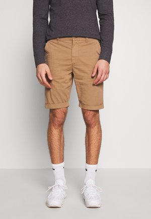 Homme portant un short beige retroussé, des chaussettes blanches de sport, des baskets blanches et une chemise à manches longues gris foncé, debout devant un fond uni.