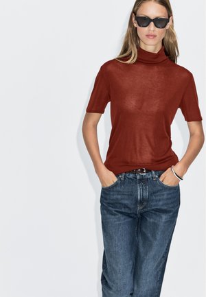 Rostfarbener, kurzärmeliger Rollkragenpullover aus leichtem Material, kombiniert mit lässigen, weiten, dunklen Jeans und einem silbernen Armband am Handgelenk.