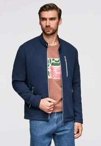 Marinblå texturerad jacka med mandarin-krage, framzipning och två sidofickor, bärs över en beigefärgad t-shirt med grafik och denimjeans.