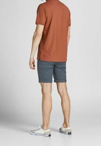 Polo shirt ruggine da uomo abbinato a pantaloni corti grigi. Il polo in cotone ha maniche corte e colletto. I pantaloni sono lunghi fino al ginocchio con orlo arrotolato.