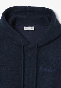 Felpa fuzzy blu navy con cappuccio e cordini, "R. Lacoste" ricamato in blu sul petto e un'etichetta Lacoste all'interno del collo.