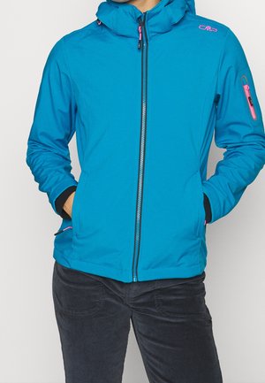 Personne portant une veste à capuche zippée bleu vif avec des détails de fermeture éclair rose et un pantalon noir, les mains dans les poches de la veste.