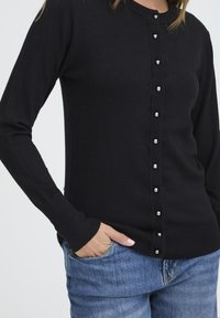 Cárdigan negro de manga larga, con cierre de botones en la parte delantera que presenta botones plateados brillantes. Puños y dobladillo acanalados, combinado con jeans azules.