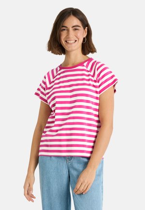 Lächelnde Frau mit kurzen braunen Haaren, die Creolen, ein rosa-weiß gestreiftes kurzärmeliges Top und hellblaue Jeans trägt und vor weißem Hintergrund steht.