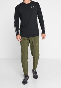 Mustat pitkät hihaiset Nike-paita, jossa on logo, yhdistettynä oliivinvihreisiin kapeisiin housuihin, joissa on logo. Mustat urheilukengät täydentävät asukokonaisuuden.
