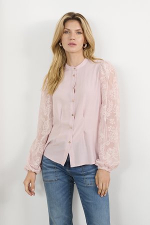 Helles rosa Bluse mit Knopfleiste, die durchscheinende, gestickte lange Ärmel und geraffte Bündchen hat. Kombiniert mit blauen Jeans.