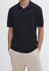 Tummansininen, lyhythihainen poolopaita, jossa on tasainen kaulus ja rento istuvuus. Paidassa on sileä pinta ja resorireunus, ja se on yhdistetty vaaleanbeigeihin housuihin.