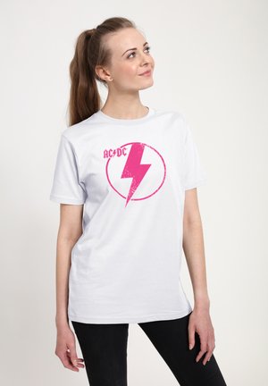 Ung kvinde med langt hestehale iført hvid T-shirt med en lyserød lyn og "AC/DC"-logo, der kigger opad på ensfarvet baggrund.