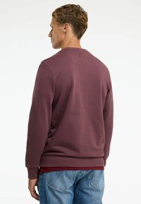 Burgundy színű pulóver bordás anyagból, hosszú ujjakkal, kerek nyakkivágással és egy kis téglalap alakú díszítéssel a háton. Kék farmernadrággal viselve.