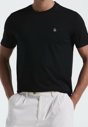 Uomo che indossa una t-shirt nera semplice a maniche corte con un piccolo logo di pinguino bianco sul petto e pantaloni bianchi plissettati con le mani in tasca.