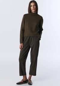 Kobieta stoi z rękami w kieszeniach, ubrana w brązowy sweter typu golf, dopasowane luźne spodnie i czarne płaskie buty z ćwiekami.