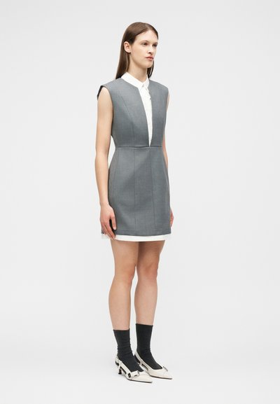 Γυναίκα όρθια, φοράει γκρι φόρεμα χωρίς μανίκια πάνω από λευκό πουκάμισο, μαύρες κάλτσες και λευκά παπούτσια με τακούνι και ασημένια αγκράφα.