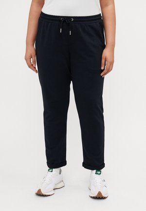 Person wearing black drawstring pants and white sneakers standing on a white background, cropped above the waist.