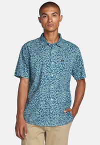 Camisa de botones de manga corta en azul con un diseño de estampado de leopardo. Confeccionada en tejido ligero, cuenta con un collar y un bolsillo con un logo.