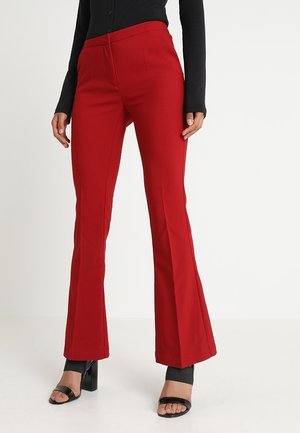 Femme portant un pantalon évasé rouge taille haute, associé à des sandales à talons noires ouvertes et un haut noir ajusté.