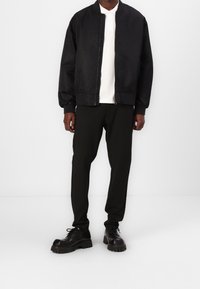 Veste bomber noire avec une fermeture éclair à l'avant, chemise blanche à col en dessous, et pantalon à rayures noires texturé. Des chaussures noires massives complètent le look.