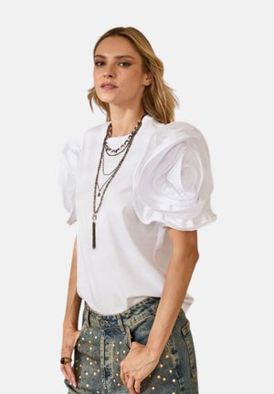 Donna con top bianco dalle grandi maniche a sbuffo arricciate e collane a catena metallica stratificate, abbinato a jeans di denim borchiati.