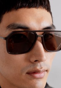 Brūni taisnstūra saulesbrilles ar metāla akcentu, tumšiem objektīviem un elegantu rāmi. Klanjot tuvplānā uz seju, neitrālā fonā.