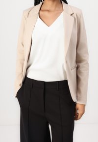 Blazer beige avec une coupe ajustée, manches longues, et un détail bouton unique, associé à un haut blanc à col en V et un pantalon noir ajusté.