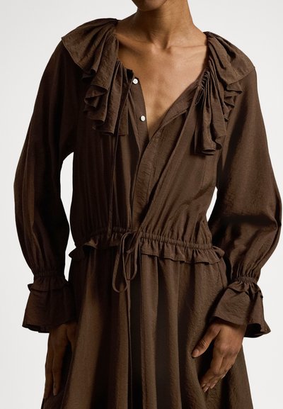 Ruda ilgomis rankovėmis suknelė su raukiniais ant apykaklės ir rankogalių. Turi raištelį juosmenyje ir užsegimą priekyje su sagomis, pagaminta iš minkštos, tekstūruotos medžiagos.