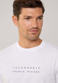 Witte katoenen T-shirt met een ronde hals en centreren marineblauwe tekst met de opdruk "FAÇONNABLE FRENCH RIVIERA." Eenvoudig, klassiek ontwerp.