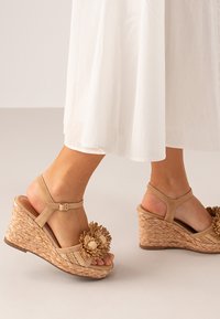 Sandalias de cuña con base de paja trenzada, correas de cuero marrón y un acento floral decorativo en la punta. Materiales texturizados y de colores claros.