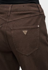 Καφέ τζιν από τζιν ύφασμα με τρίγωνο λογότυπο Guess στην πίσω τσέπη. Διαθέτουν ομαλή υφή και κλασικό σχέδιο με πέντε τσέπες.