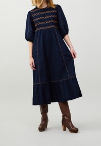 Navyblaues Midi-Kleid mit Puffärmeln, gesmocktem Oberteil und kontrastierender orangefarbener Naht. Mit einem gestuften Rüsche-Saum und glatter Stofftextur.