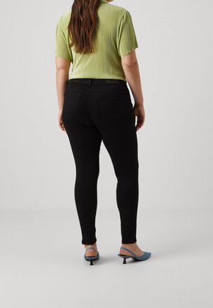 Woman with long brown hair wearing a lime green short-sleeve top, black fitted pants, and blue low-heel slingback shoes, facing away.