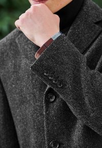 Mörkgrå ullblazer med ett texturerat herringbone-mönster, med fyra svarta knappar och en rutmönstrad skjortärm synlig under ärmen.