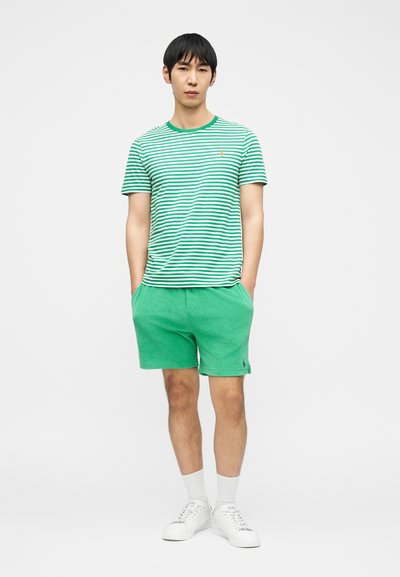 Νέος άνδρας που στέκεται με τα χέρια στις τσέπες, φοράει πράσινο και άσπρο ριγέ T-shirt, πράσινες βερμούδες, άσπρες κάλτσες και άσπρα αθλητικά παπούτσια.