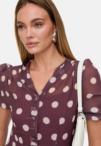 Lila Bluse mit weißen Punkten, V-Ausschnitt, kurzen Puffärmeln und Knopfverschluss. Kombiniert mit einer weißen Handtasche und großen silbernen Creolen.