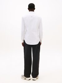 Camicia bianca con colletto e maniche lunghe, texture semplice, etichetta sul retro, abbinata a pantaloni neri a gamba larga e sneakers grigie.