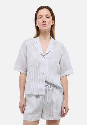 Femme portant une chemise à manches courtes gris clair avec boutons et un short assorti à cordon, avec des rayures verticales subtiles, debout devant un fond uni.