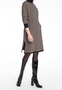 Robe en tricot avec un motif pied-de-poule beige et noir, dotée d'un col rond, de manches longues et d'entailles sur les côtés, assortie à des bottes noires brillantes jusqu'aux genoux.
