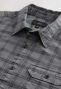 Camicia a quadri grigia con bottoni, realizzata in tessuto morbido. Presenta un colletto, bottoni neri e una tasca sul petto. Etichetta del marchio visibile.