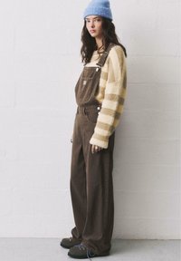 Braune Corduroy-Latzhose über einem creme- und gelb gestreiften Pullover, kombiniert mit einer blauen Mütze und dunkelgrünen Schuhen mit lila Schnürsenkeln.