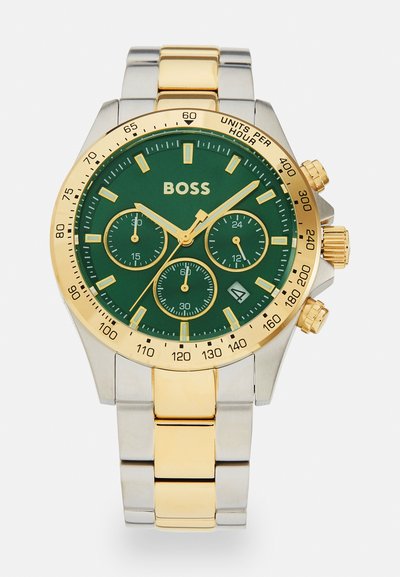 Montre avec cadran vert et accents dorés, bande en acier inoxydable et ton or, fonctionnalités chronographe et affichage de la date à la position 4 heures.
