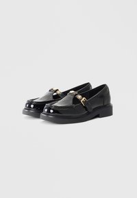 Black patent leather loafers with a smooth finish, featuring gold hardware accents and a thick, textured black sole.