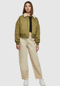 Giacca bomber verde oliva corta con colletto in eco-pelliccia crema, polsini a coste, zip frontale e tasche laterali, abbinata a pantaloni beige a gamba larga.