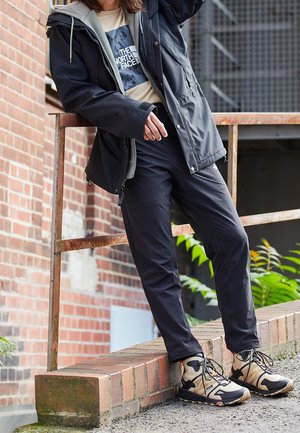Person iført sort jakke, beige The North Face-shirt, sorte bukser og beige vandresko, der står på en murstensafsats ved siden af et metalgelænder.