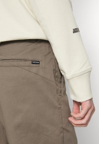 Chemise beige à manches longues avec des poignets côtelés, associée à un pantalon marron clair arborant un logo Volcom visible sur la poche arrière et une texture lisse.